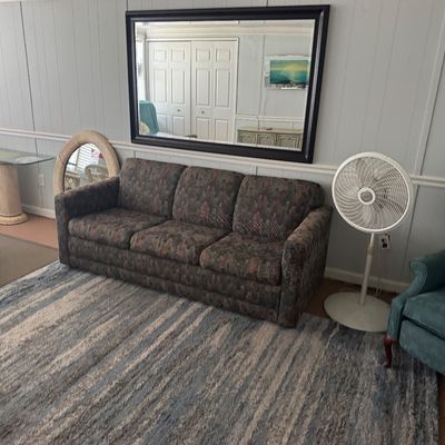 Living room set featuring a pattern-upholstered three-cushion sofa, a blue wingback recliner, a white Lasko pedestal fan, and a large...