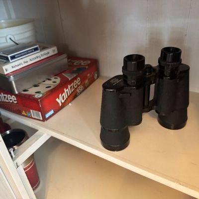 Black vintage-style binoculars displayed on a white shelf alongside a vintage Yahtzee board game and books.