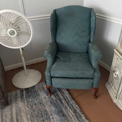 Blue flame-stitch upholstered wingback recliner with cabriole wooden front legs, paired with a white pedestal floor fan.