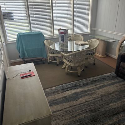 Sunroom dining set featuring a round glass-top table with four white woven rattan rolling armchairs, alongside a distressed wooden...