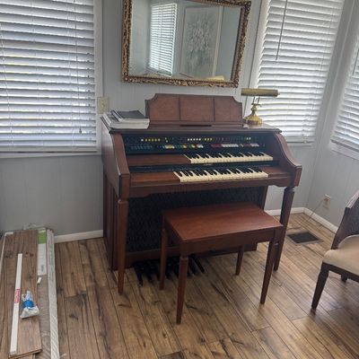 Vintage Lowrey electronic organ with double keyboard, bass pedals, and matching wooden bench, positioned beneath an ornate gold-framed...