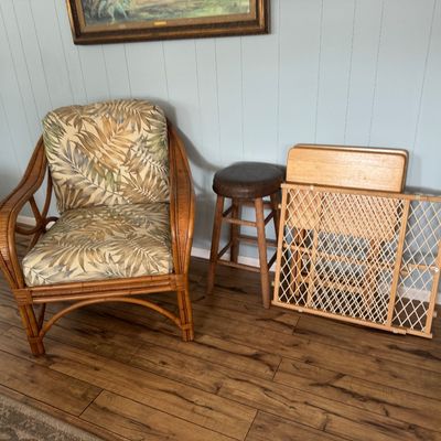 Rattan armchair with tropical leaf patterned cushions, standing alongside a wooden barstool, folding TV trays, and a wooden safety gate.