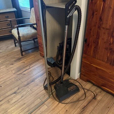 Vintage canister vacuum cleaner with a metal wand, power nozzle attachment, and long hose, stored near a mirrored closet.