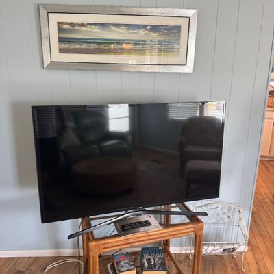 Samsung flat-screen television on a two-tier rattan stand, featuring a framed ocean landscape print mounted above.