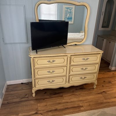 French Provincial style double dresser with an attached ornate mirror, finished in cream paint with gold trim accents, accompanied by a...