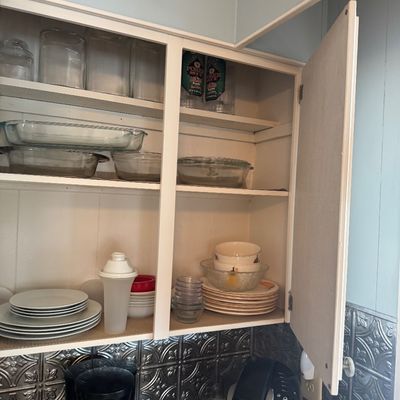 Open kitchen cabinet containing assorted glassware, Pyrex baking dishes, ceramic plates, and bowls, situated above a countertop with a...