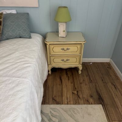 French Provincial style two-drawer nightstand in cream paint with gold trim, topped with a small cottage-style table lamp.
