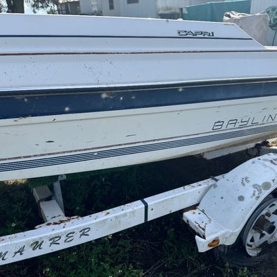 Side profile of a Bayliner Capri powerboat resting on a weathered trailer, showing some surface wear and age.