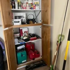 Sale Photo Thumbnail #34: Tall pine storage cabinet filled with household cleaning supplies, small electronics, gas cans, and a hydraulic floor jack, alongside various lawn tools.