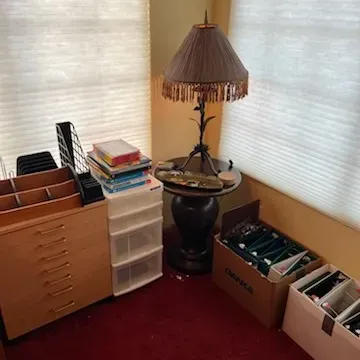 Sale Photo Thumbnail #15: Home office corner featuring a wooden accent table with a fringe-trimmed lamp, a light wood filing cabinet, a plastic storage cart, and boxes of organized files.