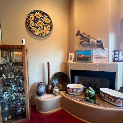 Sale Photo Thumbnail #2: Living room area featuring a fireplace hearth with ceramic vessels, a taxidermy fish mount, framed Southwestern artwork, a decorative wall plate, and a wooden glass-door display cabinet filled with figurines.