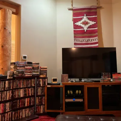 Sale Photo Thumbnail #48: Home entertainment setup featuring a flat-screen TV on a wood media console, extensive DVD and Blu-ray media collection on shelves, a Southwest-style woven wall hanging, and a tufted leather ottoman.