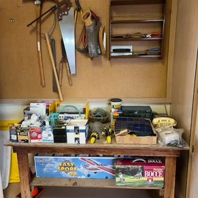 Sale Photo Thumbnail #42: A wooden workbench arranged with various hand tools, lawn games, model airplane kits, hardware, and accessories, topped with a wall-mounted pegboard holding vintage saws and garden tools.