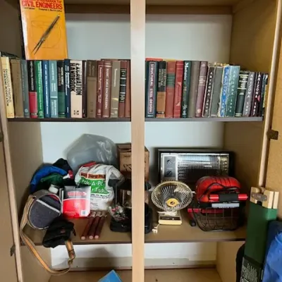 Sale Photo Thumbnail #41: A light wood storage cabinet filled with engineering handbooks and textbooks on the top shelf, above various household supplies, small electric heaters, and fans.