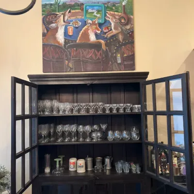 Sale Photo Thumbnail #23: Dark wood display cabinet filled with an assortment of glassware and steins, topped with a whimsical desert-themed coyotes canvas painting.
