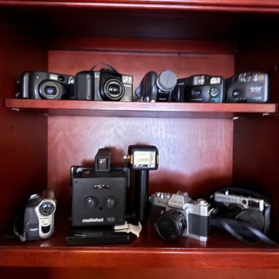 Sale Photo Thumbnail #76: Collection of vintage cameras and camcorders, including point-and-shoot models, a Canon 35mm SLR, a Polaroid Miniportrait, and digital video cameras, displayed on wooden shelves.