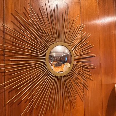 Sale Photo Thumbnail #111: Mid-century modern sunburst wall mirror featuring a gold-finished frame with radiating metal rods, mounted on wood paneling.