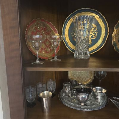 Sale Photo Thumbnail #211: Collection of vintage decorative glassware, silver-tone tea and cream service pieces, and gilt Florentine-style trays displayed in a wooden cabinet.