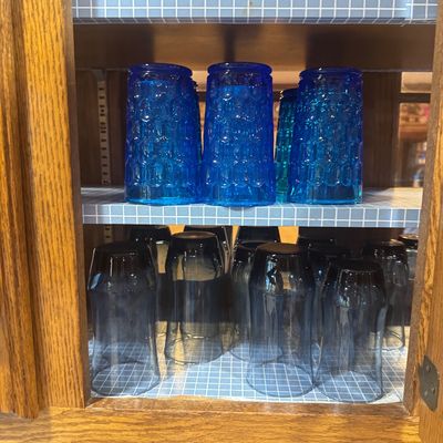 Sale Photo Thumbnail #223: Collection of vintage glass drinking tumblers displayed inside a wooden cabinet, featuring cobalt blue textured glasses on the top shelf and dark smoke blue glasses below.