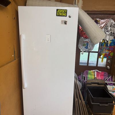Sale Photo Thumbnail #94: White upright freezer with a front control panel and top stickers, shown stored in a garage alongside folding chairs and household items.