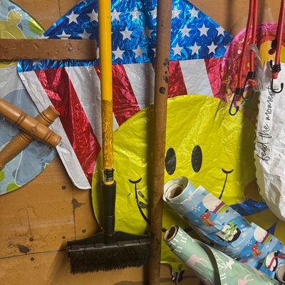 Sale Photo Thumbnail #93: An assortment of household items stored against a particle board wall, including deflated Mylar balloons, long-handled cleaning tools, and rolls of holiday wrapping paper.