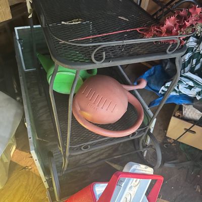 Sale Photo Thumbnail #2: Black wrought iron outdoor rolling bar cart with mesh tiers, displayed alongside plastic watering cans and a vintage red Coleman cooler.