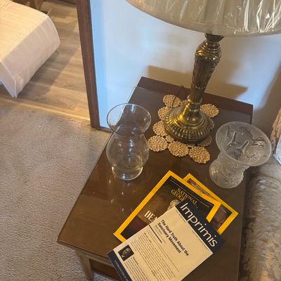 Sale Photo Thumbnail #168: Vintage brass table lamp with a plastic-wrapped shade resting on a crochet doily alongside two glass vases and National Geographic magazines on a wooden end table.