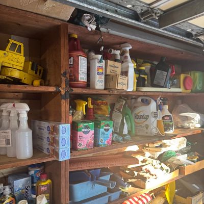 Sale Photo Thumbnail #82: Wooden garage shelving unit filled with household supplies, vintage yellow Tonka construction toy, cleaning chemicals, garden products, and miscellaneous hand tools.