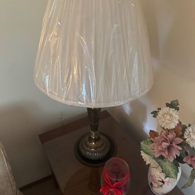 Sale Photo Thumbnail #149: Vintage brass-tone table lamp featuring a pleated white lampshade wrapped in protective plastic, displayed on a wooden side table.