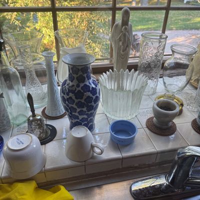 Sale Photo Thumbnail #229: Collection of glass and ceramic items lined up on a windowsill, including a blue and white floral vase, clear glass vases, white milk glass, and a small Madonna figurine.