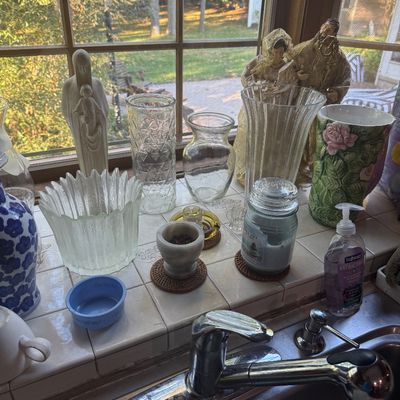 Sale Photo Thumbnail #226: Collection of glass vases, religious figurines, and household items arranged on a tiled windowsill above a kitchen sink.