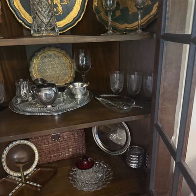 Sale Photo Thumbnail #209: Vintage glass and silver-plated tableware displayed in a wooden cabinet, including etched stemware, cut-crystal vases, decorative trays, and cream and sugar servers.