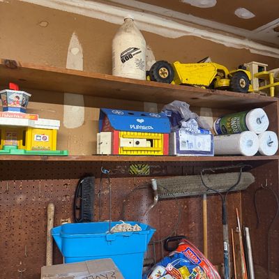 Sale Photo Thumbnail #91: Workshop shelves featuring vintage toys including a Tonka construction vehicle and a Playskool wooden barn, along with cleaning supplies and garden tools.