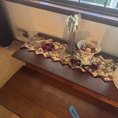 Sale Photo Thumbnail #186: Dark wood console table styled with a quilted table runner, a silver-tone angel statue, and a decorative white pierced ceramic bowl.
