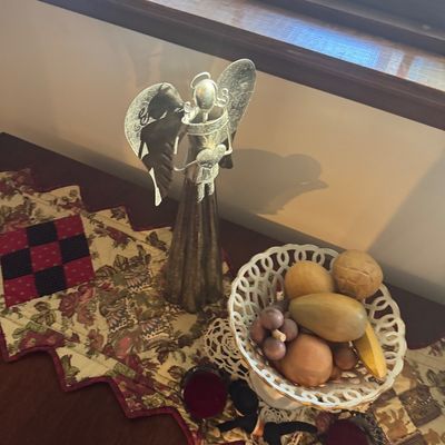 Sale Photo Thumbnail #158: A silver-toned metal angel figurine standing beside a white reticulated ceramic fruit bowl filled with decorative wooden fruit, set atop a patchwork quilted runner.