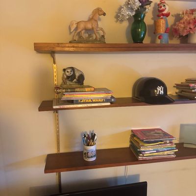 Sale Photo Thumbnail #56: Wall-mounted wood shelving unit filled with decor, including figurine collectibles, books, a green glass vase with flowers, and a New York Yankees baseball helmet.