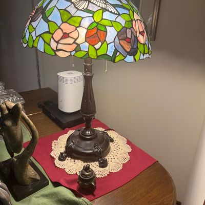 Sale Photo Thumbnail #196: Tiffany-style table lamp featuring a colorful stained glass shade with hummingbird and floral motifs over a dark bronze-finished base.