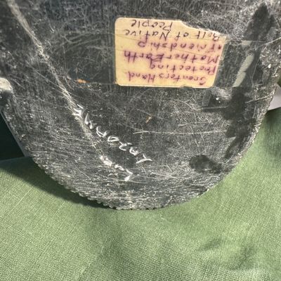 Sale Photo Thumbnail #192: Underside of an Inuit soapstone carving featuring an etched artist signature and a handwritten taped label titled 'Creator's Hand Protecting Mother Earth'.
