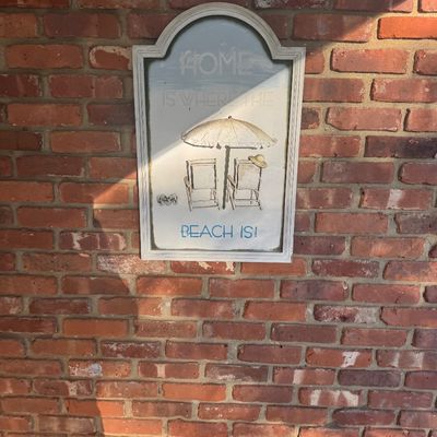 Sale Photo Thumbnail #112: Framed coastal wall art piece with an arched top, featuring beach chairs, an umbrella, and the text 'Home is where the beach is!' mounted on a red brick wall.