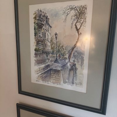Sale Photo Thumbnail #175: Framed watercolor print depicting a Parisian street scene along the Seine, matted and set in a black frame.