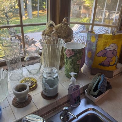 Sale Photo Thumbnail #225: Assorted household items arranged on a tiled windowsill including clear glass vases, a floral ceramic vase, religious figurines, a jarred candle, and a small mortar and pestle.