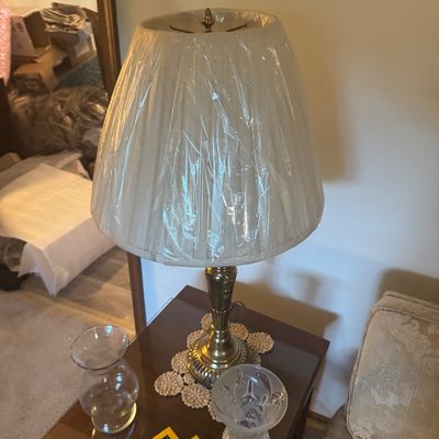 Sale Photo Thumbnail #169: Traditional brass table lamp with a plastic-wrapped pleated shade resting on a crocheted doily atop a dark wood side table.