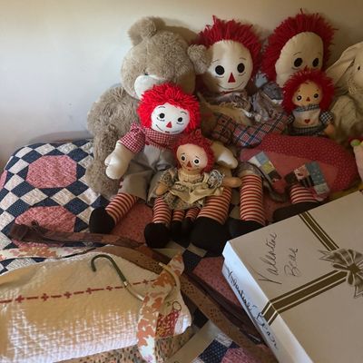 Sale Photo Thumbnail #65: Collection of Raggedy Ann and Andy style rag dolls displayed alongside plush teddy bears, vintage textiles, belts, and a labeled storage box on a patchwork quilt.