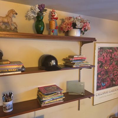 Sale Photo Thumbnail #57: Wall-mounted three-tier wood shelving unit displaying various books, a New York Yankees souvenir batting helmet, floral arrangements, decorative figurines, and a framed Callaway Gardens poster.