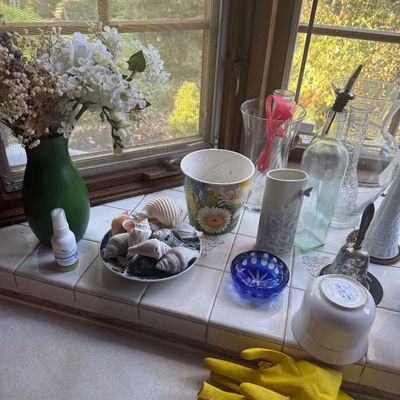 Sale Photo Thumbnail #227: A windowsill collection featuring assorted glass vases, a plate of seashells, a cobalt blue glass dish, a ceramic flower pot, and artificial floral arrangements.
