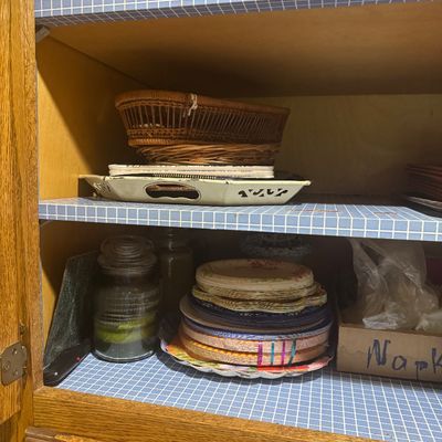 Sale Photo Thumbnail #243: Wooden cabinet shelves filled with kitchenware, including a stack of disposable paper plates, a jar candle, woven baskets, and a serving tray.