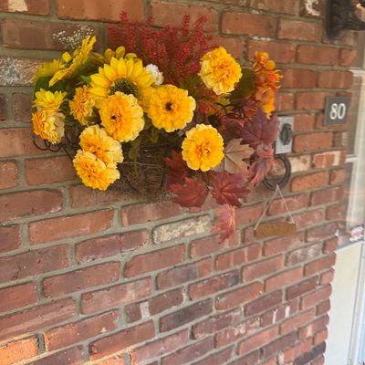 Sale Photo Thumbnail #111: Autumn artificial floral arrangement featuring yellow marigolds, sunflowers, and faux fall leaves mounted on an exterior brick wall.