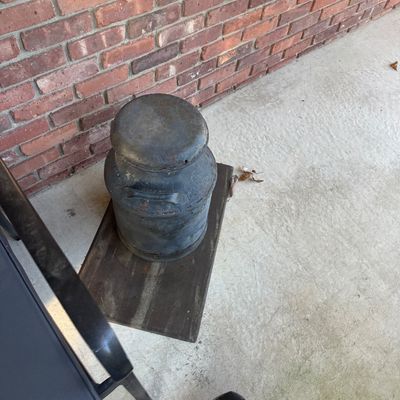 Sale Photo Thumbnail #116: Rustic vintage metal milk can with lid, sitting on a dark wooden board along a brick wall.