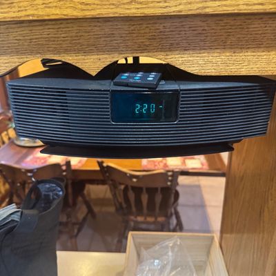 Sale Photo Thumbnail #216: Bose Wave radio mounted under a wooden shelf with remote control, featuring a digital display.