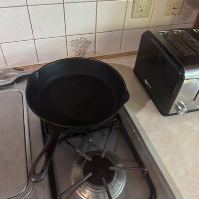 Sale Photo Thumbnail #230: Seasoned black cast iron skillet with dual pour spouts, sitting on a gas stovetop next to a toaster.